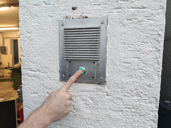 An einer weißen Rauputz Außenwand ist ein silbernes Klingelschild. Diese Klingel wird gerade von dem Zeigefinger einer Hand betätigt.