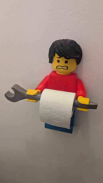 An einer weiße Wand hängt ein überdimensioniertes Klemmbausteinmännchen. Es hat schwarze Haare, einen roten Pully und eine blaue Hose an. In der Hand hält er einen Schraubenschlüssel, auf welchem eine Klopapierrolle "aufgespießt" ist.