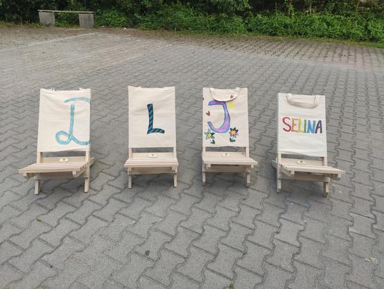 Auf gepflasterten Boden stehen 4 Liegestühle aus Holz. Auf den Stühlen wurde folgendes geschrieben (von links nach rechts): D, L, J, Selina.